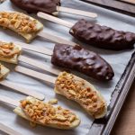 Banana pops on a baking tray with parchment paper, some are covered in chocolate, some are just topped with peanut butter and peanuts.
